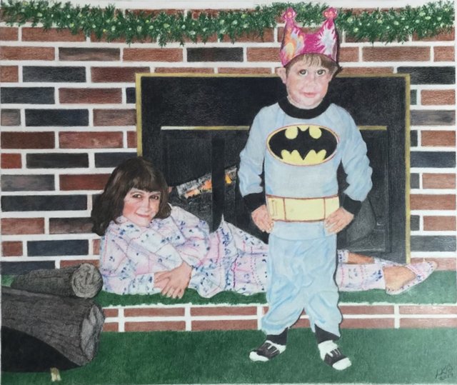 Two children in pajamas by a brick fireplace: a boy in a Batman costume and paper crown stands while a girl lounges beside him.