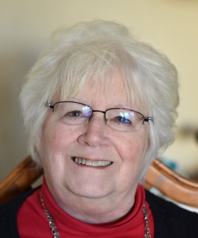 Lorna Benson, smiling, with white hair and glasses, wearing a red turtleneck.